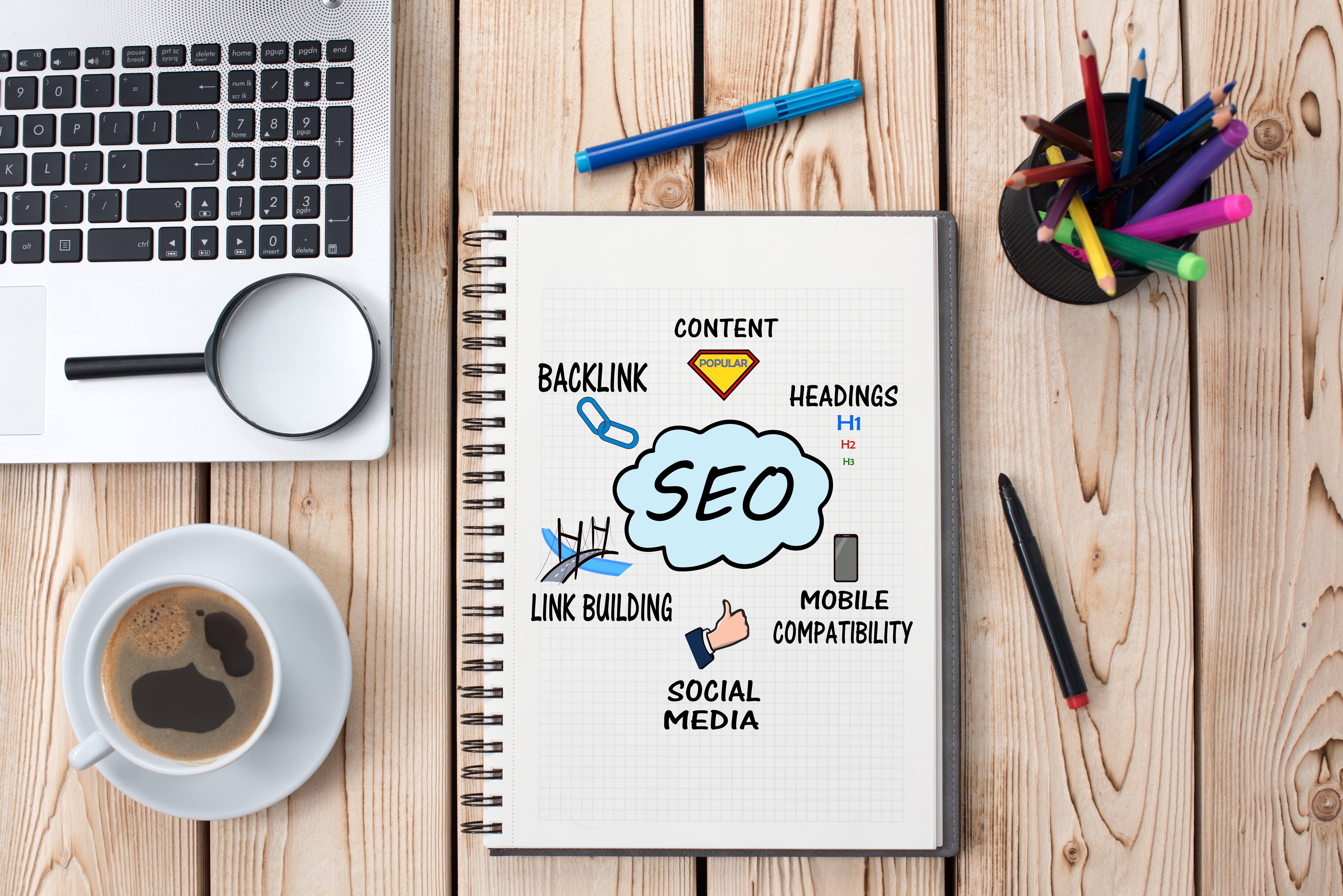 a notebook outlining SEO with content headings mobile compatability social media link building and backling. Separately ther is a cup of coffee, a loose pen and marker, a cup of pens and pencils, and a laptop keyboard with a magnifying glass resting on it