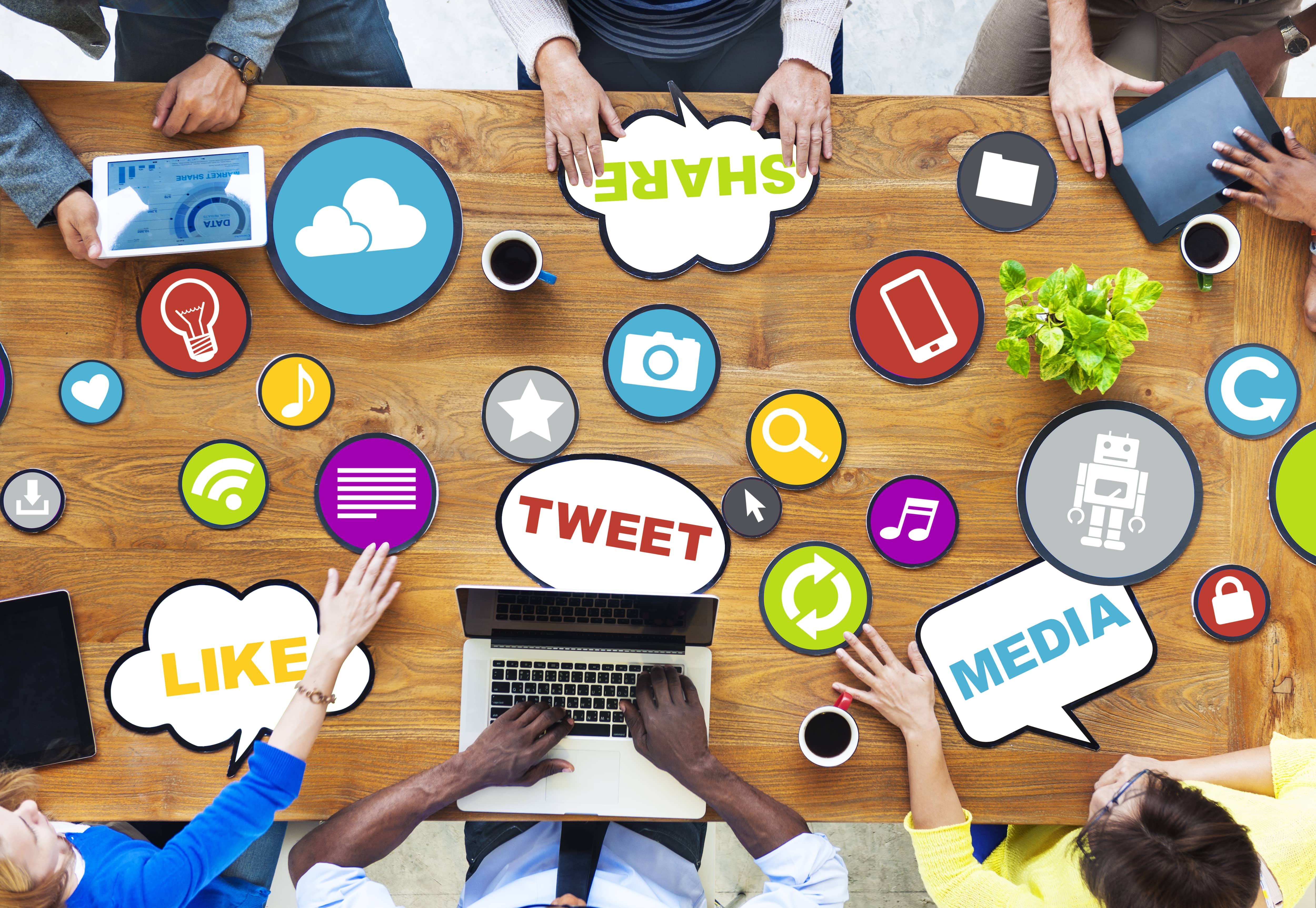 overhead veiw of a table with six people sitting at it. one person has a laptop and one person has a tablet. the rest of the people are moving buttons with various symbols relating to social media across the table while brainstorming